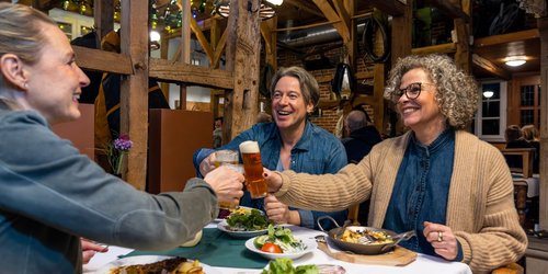 Drei Personen sitzen an einem Tisch in einem Lokal und stoßen mit Biergläsern an.
