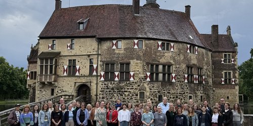 42 Personen stehen nebeneinander in zwei Reihen vor einer historischen Burg. Der Himmel ist bedeckt.