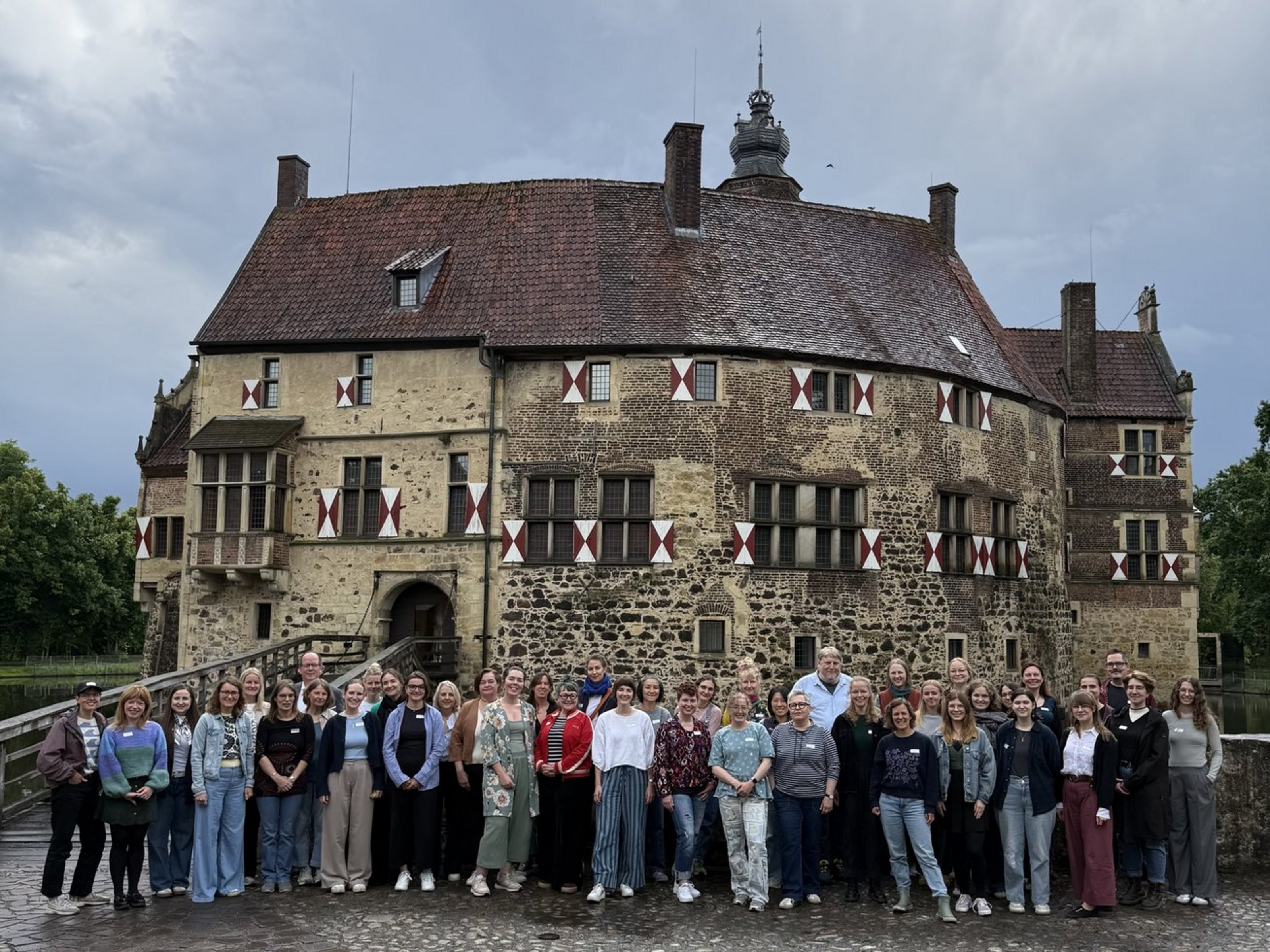 42 Personen stehen nebeneinander in zwei Reihen vor einer historischen Burg. Der Himmel ist bedeckt.