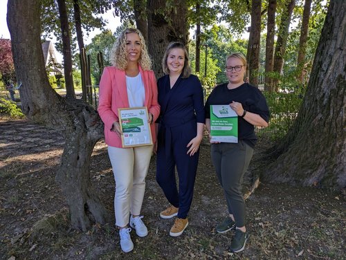 Drei Frauen stehen in einem Waldstück, zwei halten Urkunden in die Kamera.