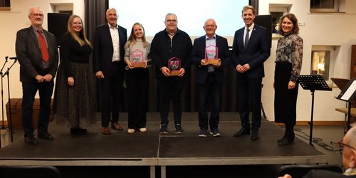 Gruppenfoto der Preisträgerinnen und Preisträger des Heimatpreises 2025 mit Laudatorinnen und Laudatoren auf einer Bühne, alle lächeln und halten Urkunden oder Trophäen in den Händen.