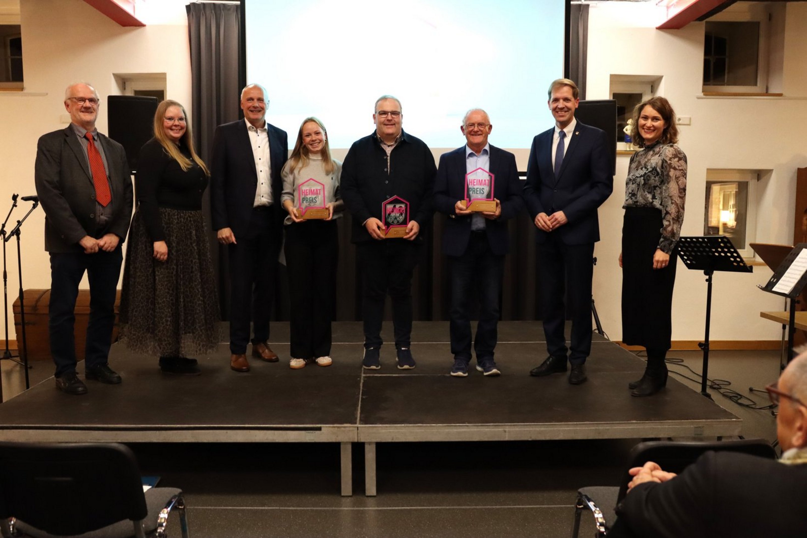 Gruppenfoto der Preisträgerinnen und Preisträger des Heimatpreises 2025 mit Laudatorinnen und Laudatoren auf einer Bühne, alle lächeln und halten Urkunden oder Trophäen in den Händen.
