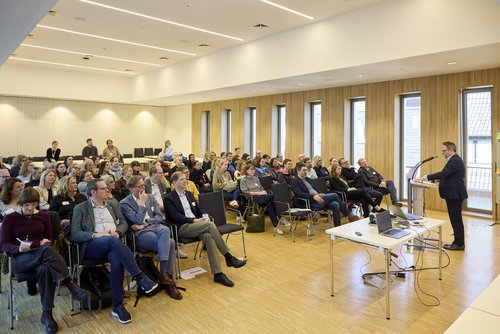 Ein Mann steht hinter einem Rednerpult vor einem Publikum aus etwa 40 Personen, welches sitzt. 