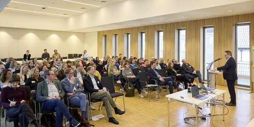 Ein Mann steht hinter einem Rednerpult vor einem Publikum aus etwa 40 Personen, welches sitzt. 