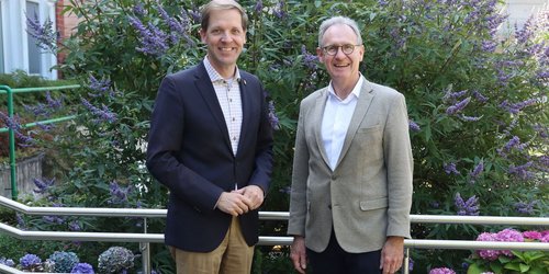 Zwei Männer in Anzug stehen vor einem Strauch mit lila Blüten und lächeln in die Kamera.