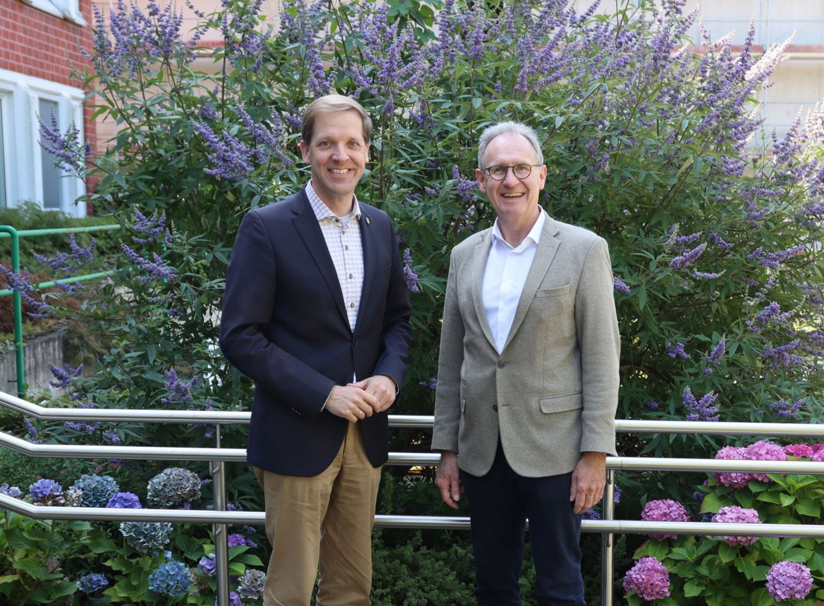 Zwei Männer in Anzug stehen vor einem Strauch mit lila Blüten und lächeln in die Kamera.