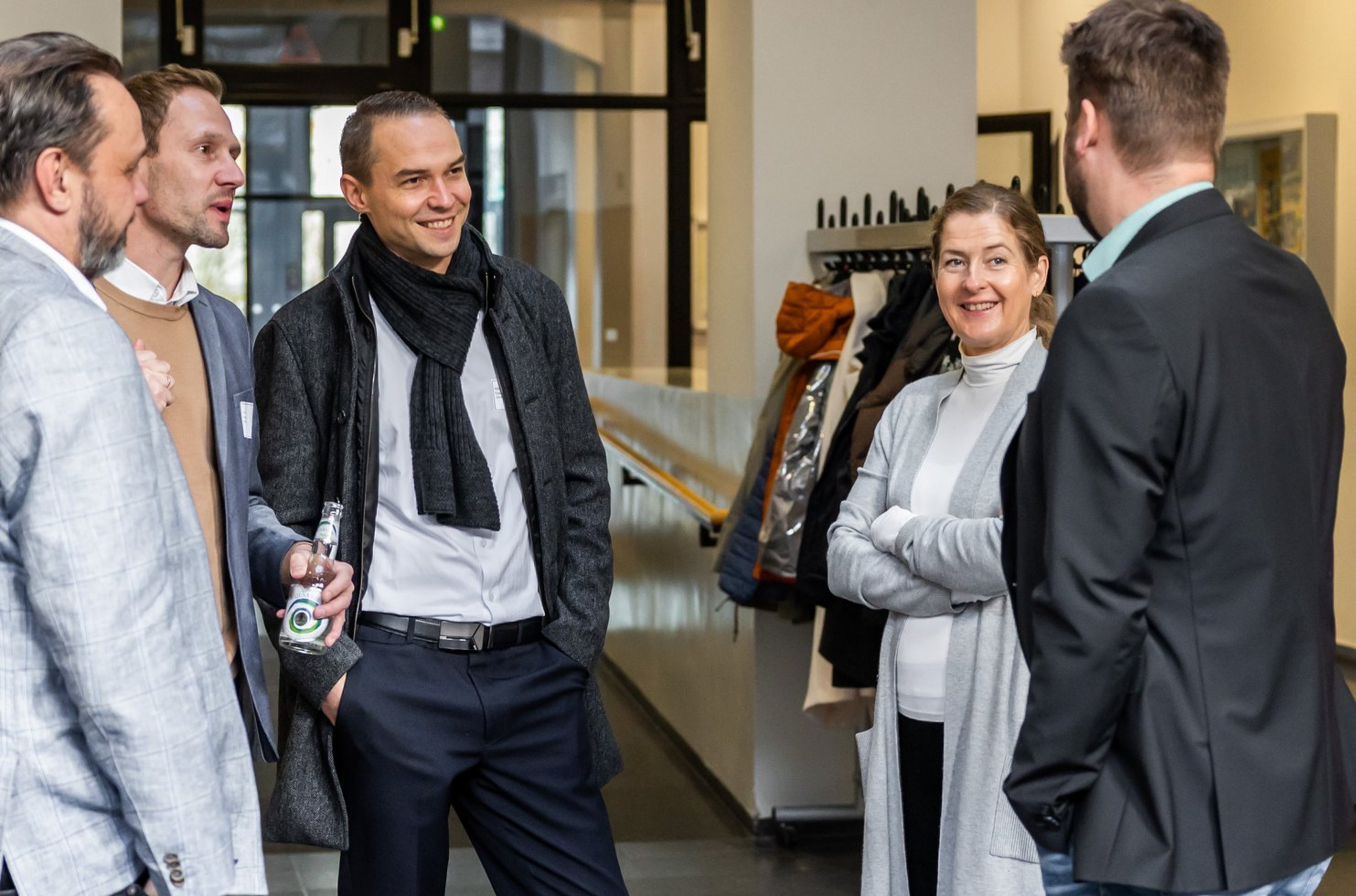 Fünf Personen in formeller Kleidung unterhalten sich in einem hellen Raum mit Garderobe im Hintergrund