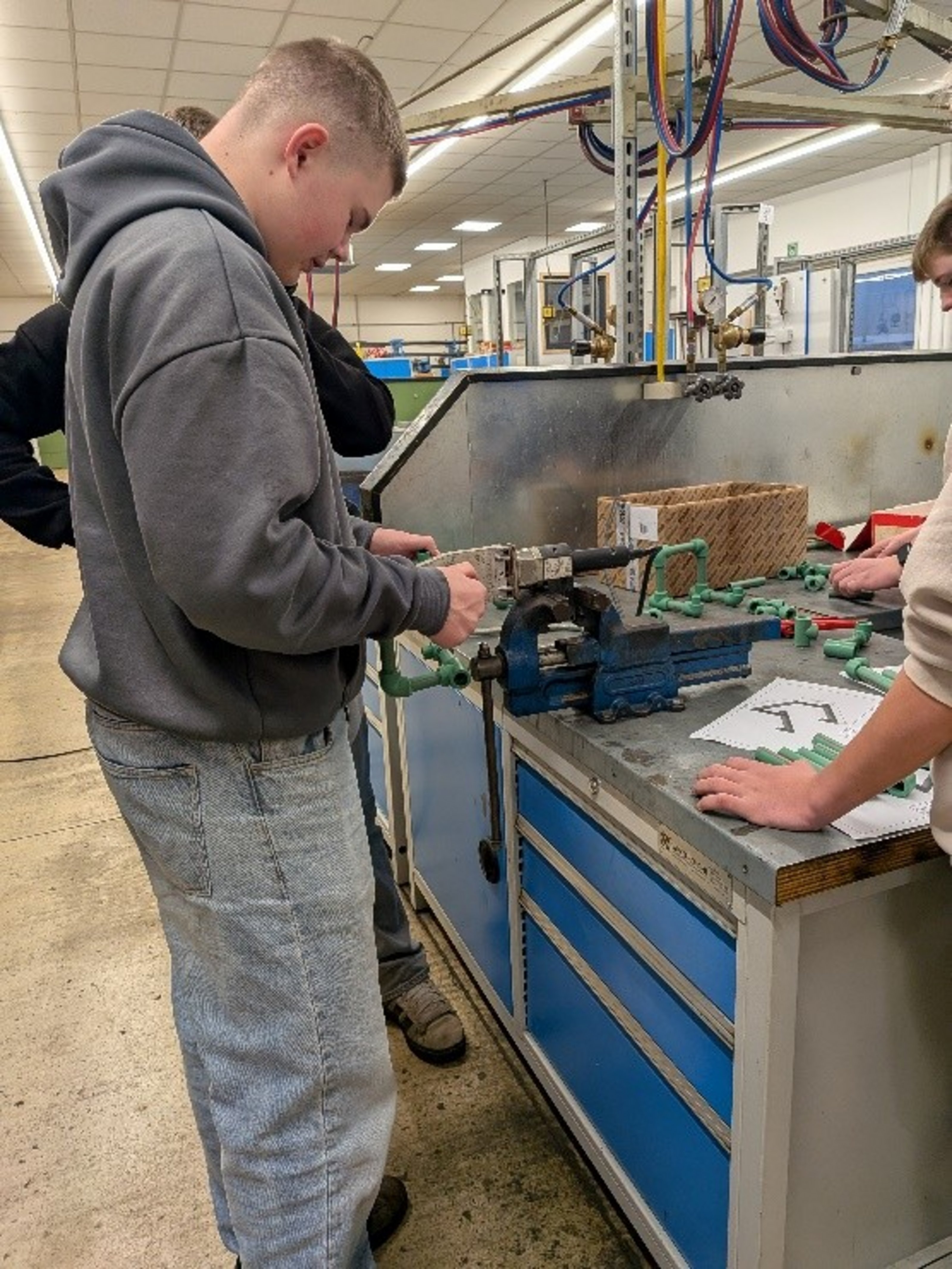 Ein Jugendlicher steht an einer Werkbank und hält ein grünes Rohr an eine Maschine.