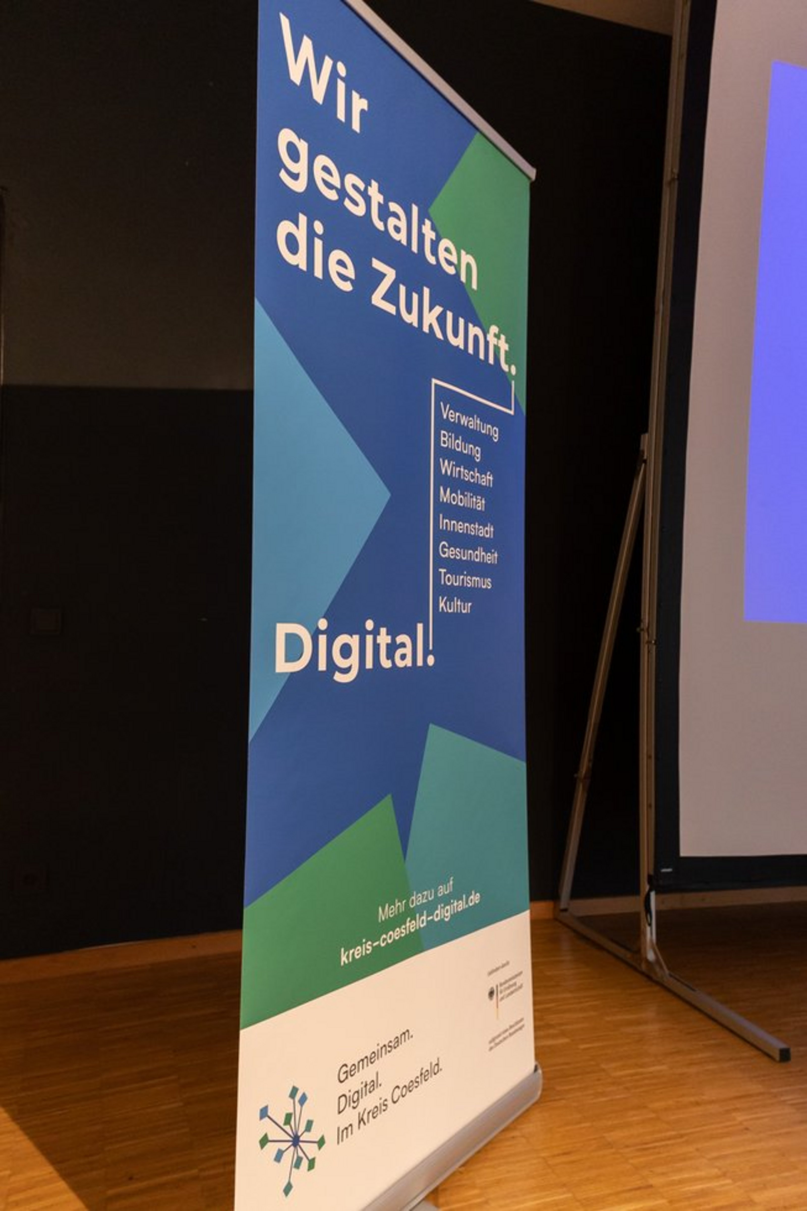 Stehendes Banner mit Text 'Wir gestalten die Zukunft Digital' und grafischem Element in einem Raum mit Leinwand