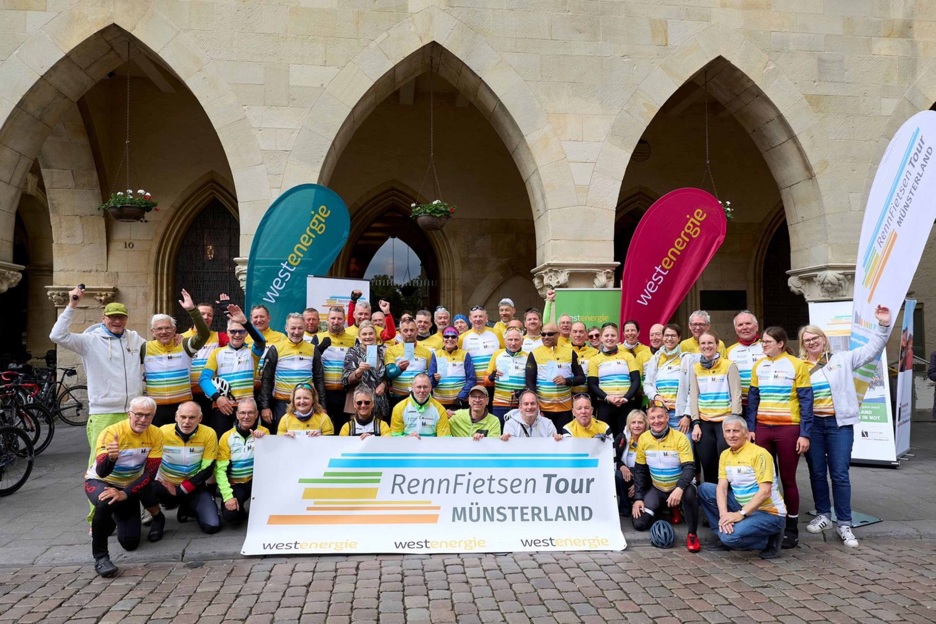 Eine große Gruppe von Radfahrerinnen und Radfahrern in gelben Trikots posiert mit einem Banner der „RennFietsen Tour Münsterland“ vor den Bögen des Prinzipalmarkts in Münster.