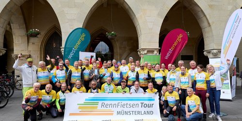 Eine große Gruppe von Radfahrerinnen und Radfahrern in gelben Trikots posiert mit einem Banner der „RennFietsen Tour Münsterland“ vor den Bögen des Prinzipalmarkts in Münster.