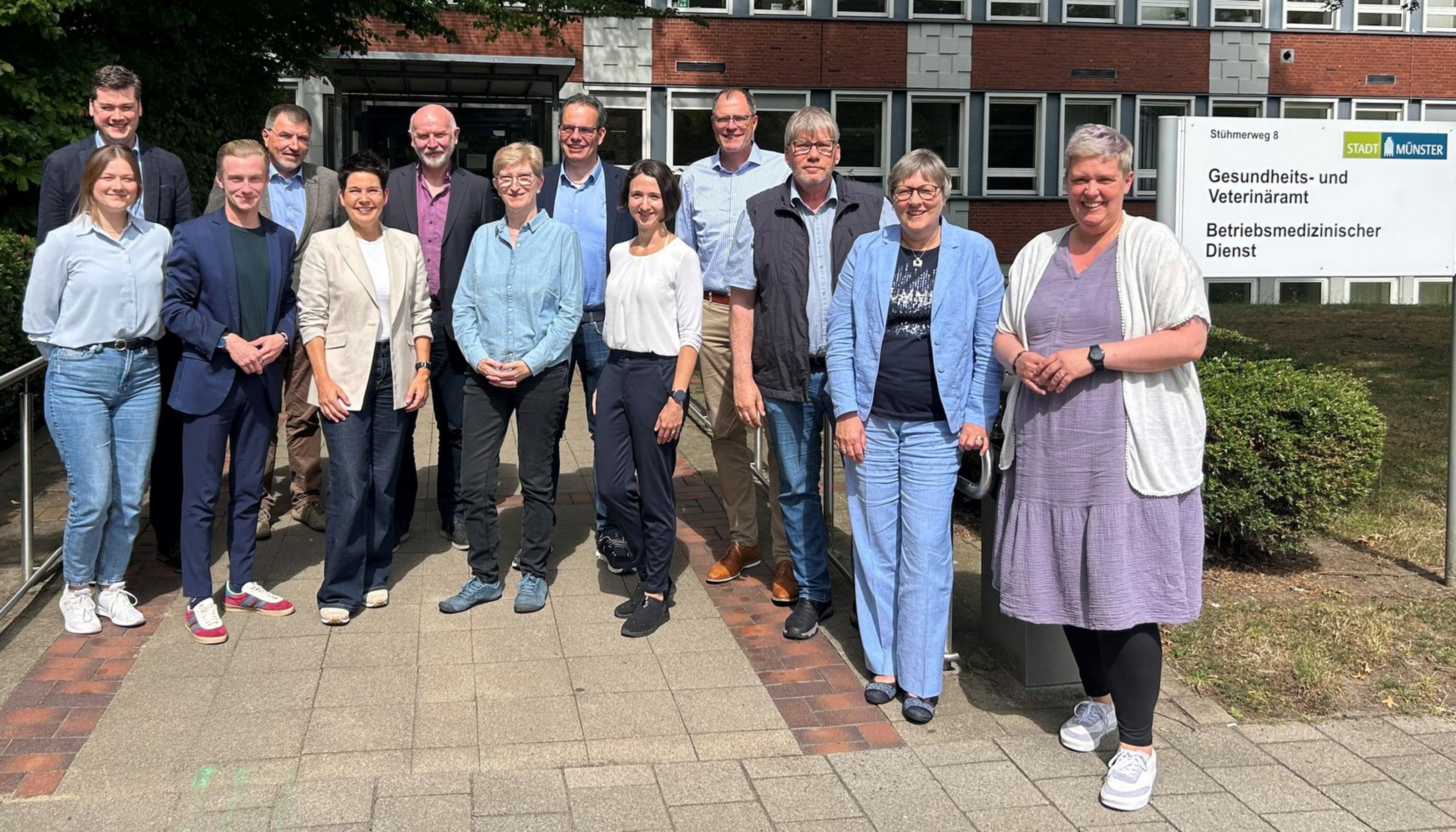 Treffen der Gesundheitsämter, des Münsterland e.V. und der KVWL in Münster (Foto: Stadt Münster). 13 Personen stehen vor einem geklinkerten Gebäude. Auf einem Schild rechts steht "Gesundheits- und Veterinäramt. Stadt Münster".