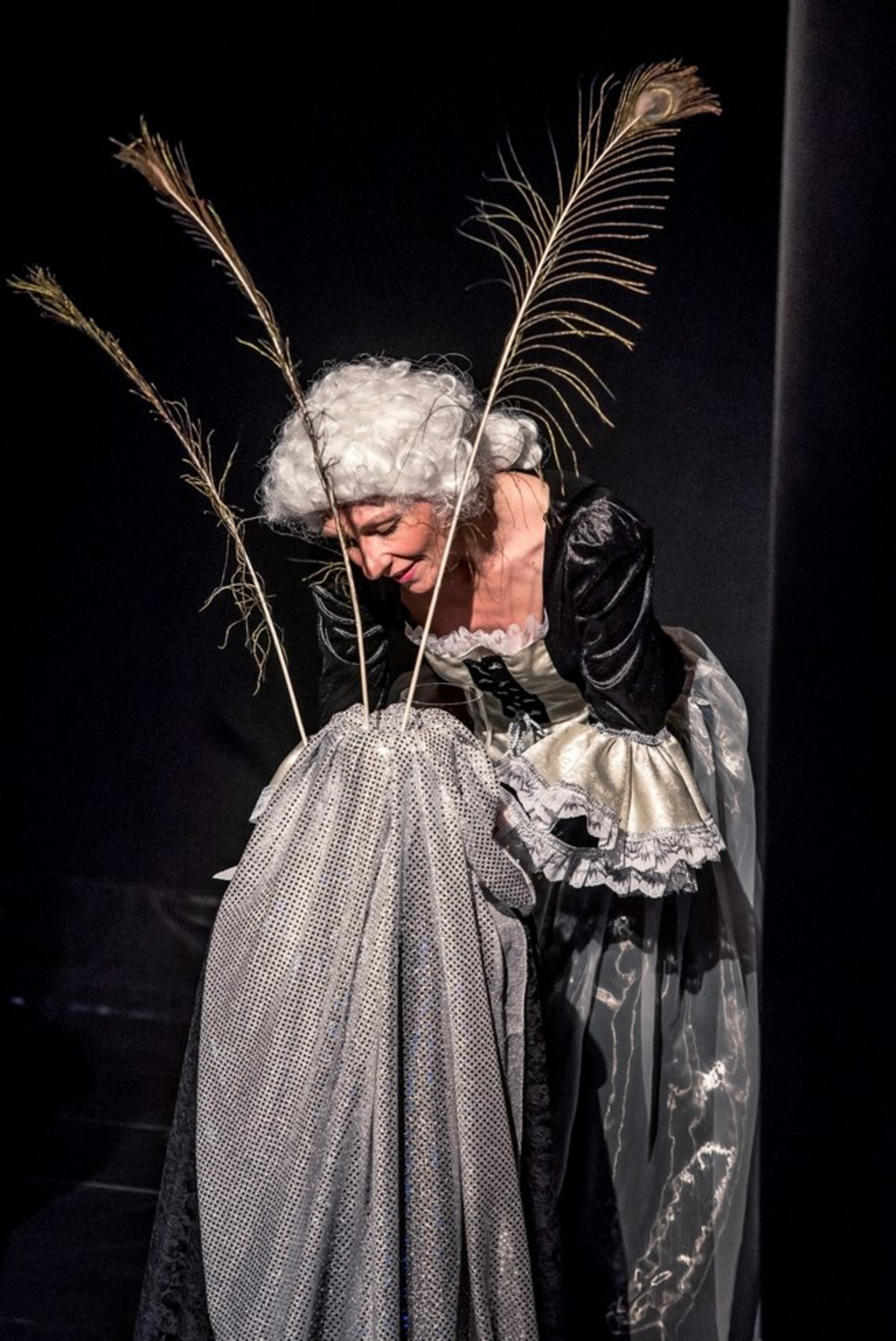 Eine Frau in historischem Kostüm und Perücke hält ein Stoff in der Hand aus dem drei Pfauenfedern ragen.