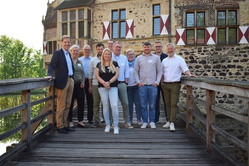 Eine Gruppe von elf Personen steht auf der hölzernen Brücke von Burg Vischering. Im Hintergrund ist das historische Gebäude mit Fachwerk und Fensterläden in Rot-Weiß. Ganz links steht Landrat Dr. Schulze Pellengahr, der lächelnd in die Kamera blickt. Neben ihm stehen die Leiterinnen und Leiter der Workshops, ebenfalls freundlich blickend. 
