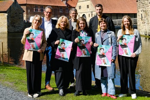 Acht Personen stehen in zwei Reihen vor dem Wassergraben eines historischen Gemäuers. Die vorderen fünf Personen halten Plakate in den Händen.
