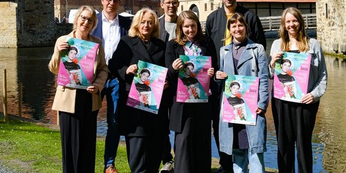 Acht Personen stehen in zwei Reihen vor dem Wassergraben eines historischen Gemäuers. Die vorderen fünf Personen halten Plakate in den Händen.