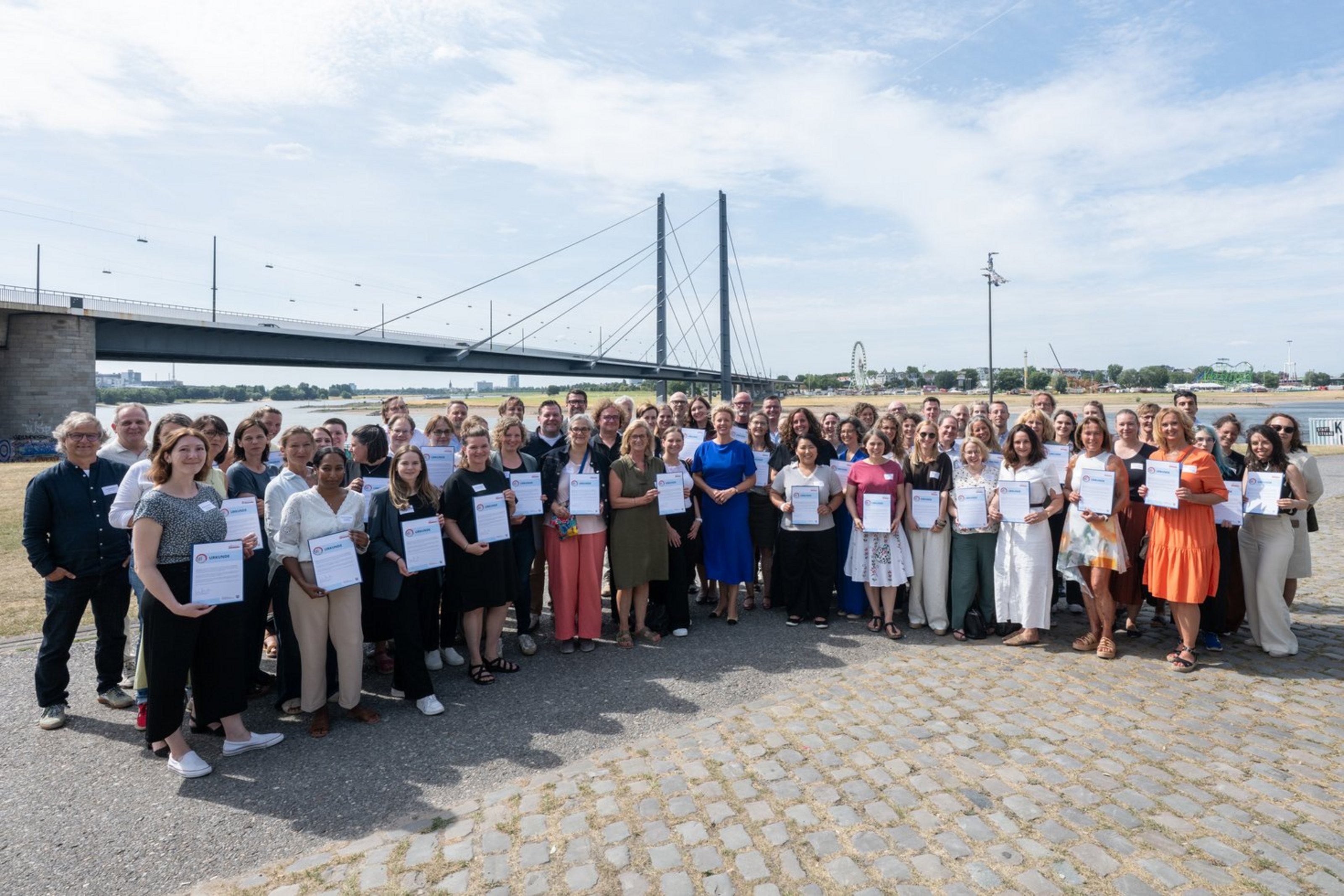 Eine große Gruppe Männer und Frauen steht vor einem Fluß, über den eine Brücke führt. Viele in der Gruppe halten ein papiernes Zertifikat in der Hand.