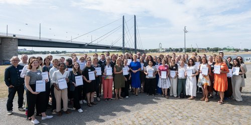 Eine große Gruppe Männer und Frauen steht vor einem Fluß, über den eine Brücke führt. Viele in der Gruppe halten ein papiernes Zertifikat in der Hand.