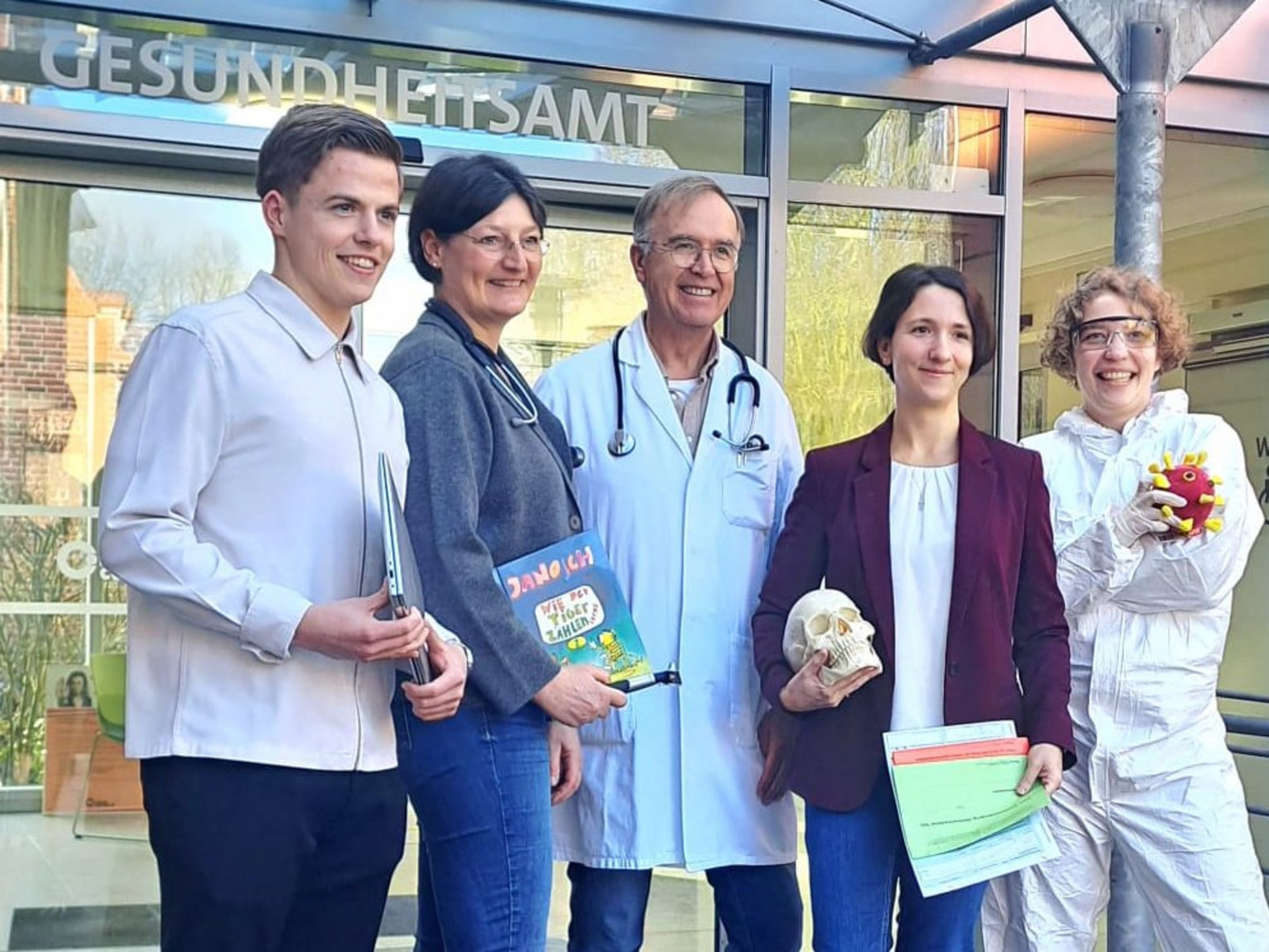 Gruppenfoto vor einem Gesundheitsamt: Fünf Mitarbeitende, drei Frauen und zwei Männer, stehen lächelnd in einer Reihen vor einem Eingangsbereich. Der Arzt in der Mitte trägt einen weißen Klittel und ein Stethoskop. Die anderen Personen haben eine nachbildung eines menschlichen Schädels, drei Bücher und eine Nachbildung eines Virus in den Händen.