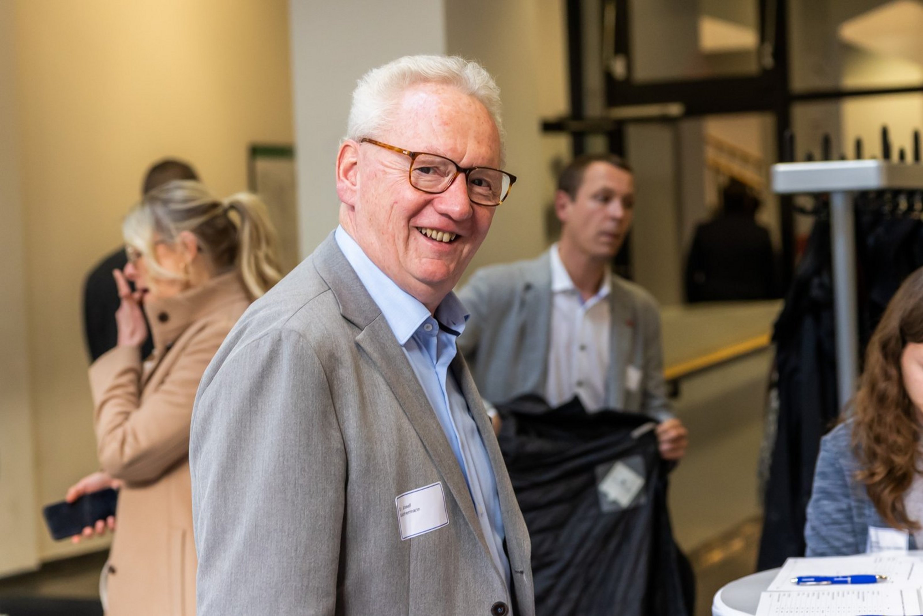 Älterer Mann mit Brille und grauem Sakko lächelnd in einem Innenraum mit mehreren Personen im Hintergrund