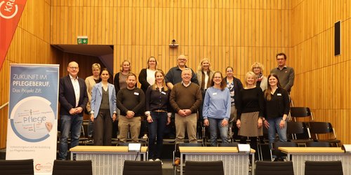 16 Personene stehen in zwei Reihen in einem Raum mit Holzvertäfelung. Sie schauen zur Kamera. Links steht ein Roll-Up mit der Aufschrift "Zukunft im Pflegeberuf".