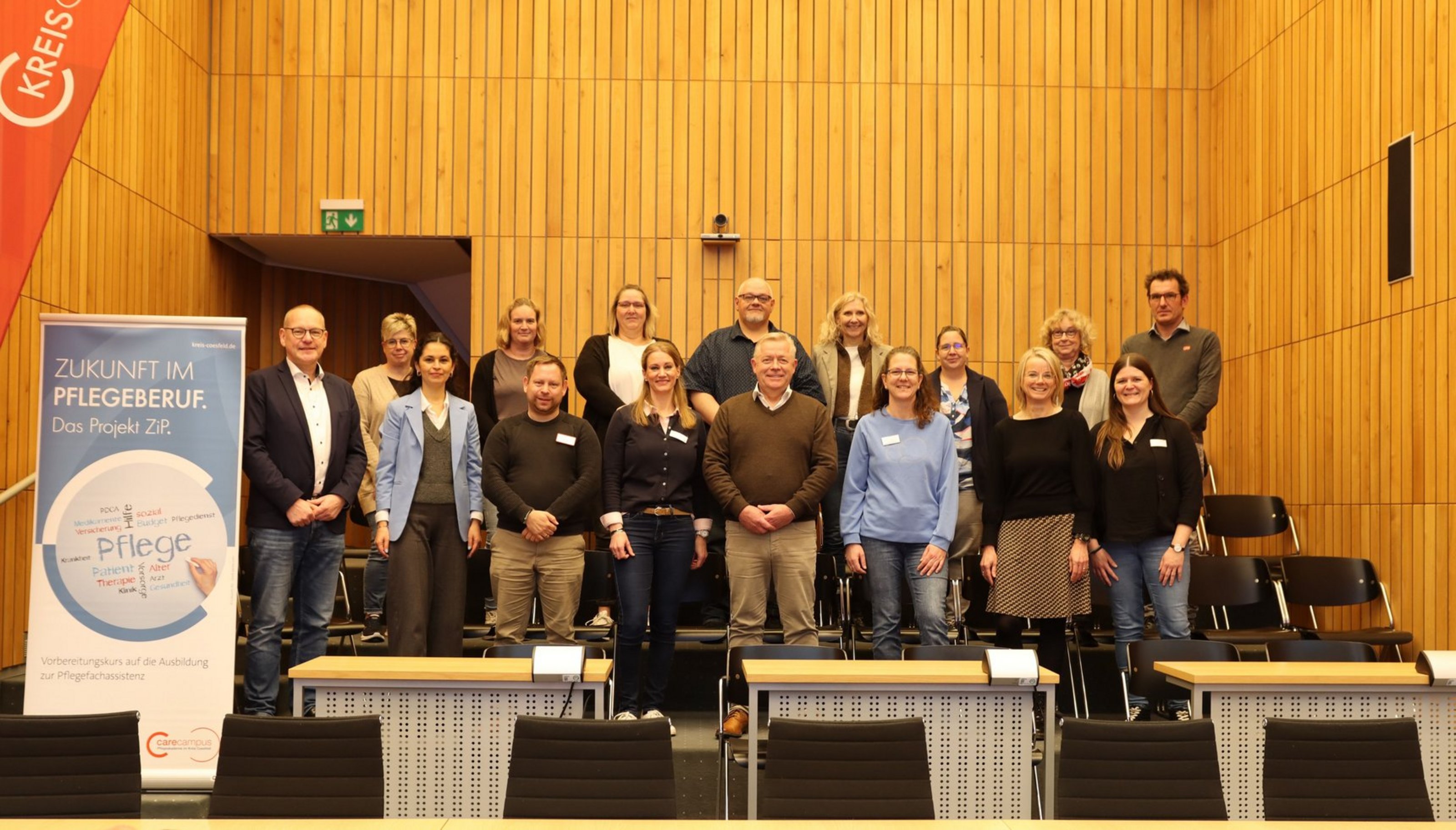 16 Personene stehen in zwei Reihen in einem Raum mit Holzvertäfelung. Sie schauen zur Kamera. Links steht ein Roll-Up mit der Aufschrift "Zukunft im Pflegeberuf".