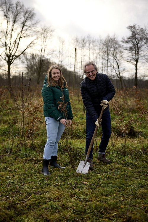 Ein Mann und eine Frau stehen auf einer Wiese. Die Frau hält einen kleinen Baum in der Hand, der Mann hält einen Spaten.