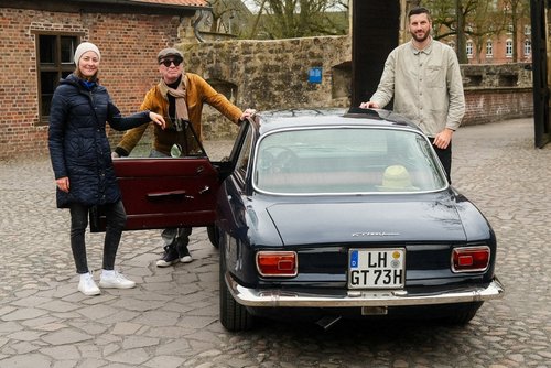 Drei Personen stehen neben einem dunkelblauen Oldtimer mit dem Kennzeichen „LH GT 73H“ auf einem historischen Pflasterweg vor einem Torbogen. Links steht eine Frau mit dunkler Jacke, Mütze und weißen Turnschuhen. In der Mitte ein älterer Mann mit brauner Jacke, Schiebermütze und Sonnenbrille, der die geöffnete Fahrertür hält. Rechts lehnt ein Mann mit heller Jacke und dunkler Hose am Dach des Autos. Im Hintergrund sind eine Backsteinmauer, ein historisches Gebäude und Bäume zu sehen. Das Organisationsteam (v.l.n.r.) aus Amelie Schulze Spüntrup, Uwe Stecker (Bertelsbeck Automotive) und Kreismitarbeiter Lukas Bertels; Kreis-Kulturreferentin Swenja Janning fehlt im Bild (Bildquelle: Kreis Coesfeld, Christoph Hüsing).