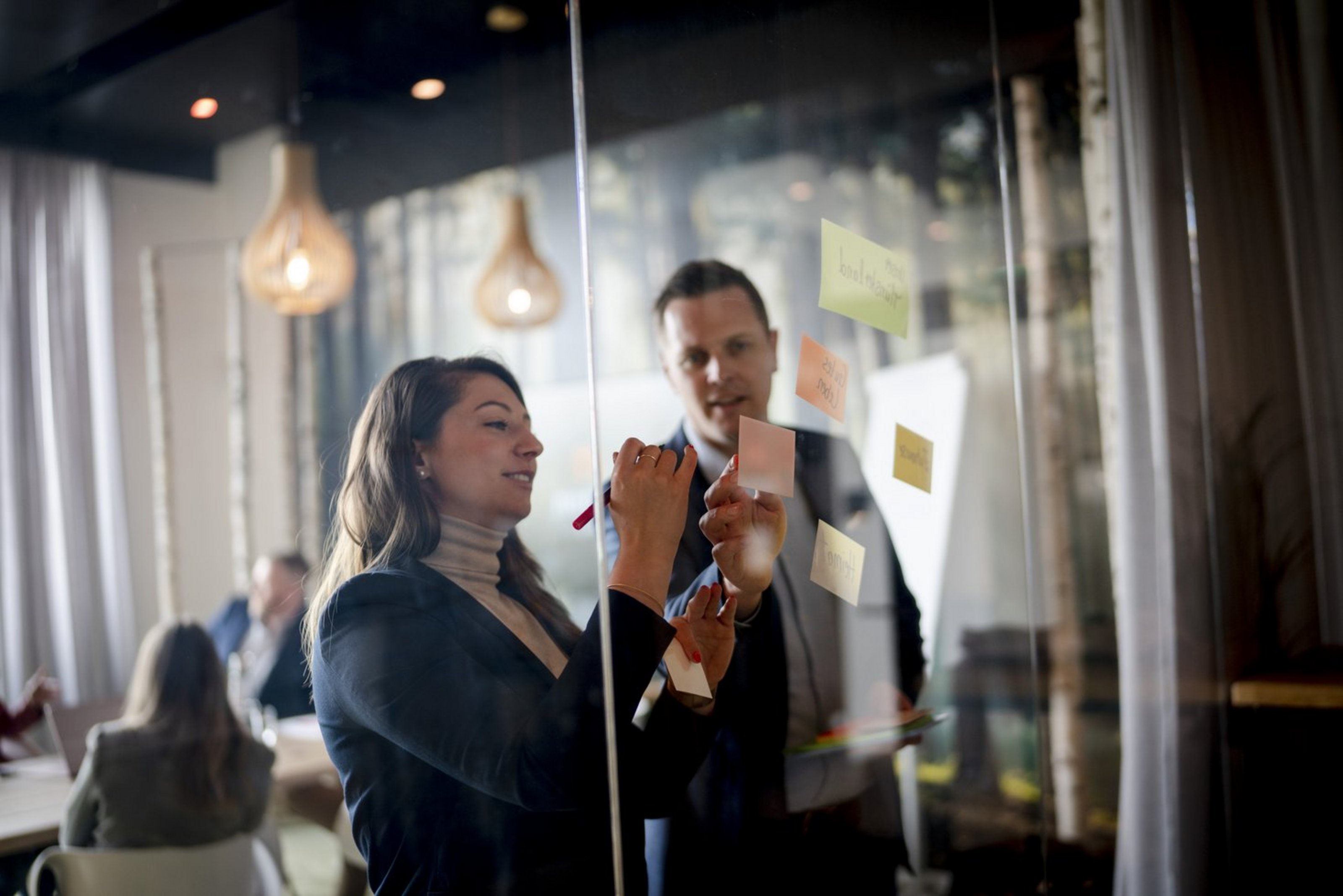 Wie es gelingt, internationale Fachkräfte fürs eigene Unternehmen im Münsterland zu begeistern, ist Thema eines Workshops für Arbeitgeberinnen und Arbeitgeber in Steinfurt. (Foto: Münsterland e.V./Anja Tiwisina) Ein Mann und eine Frau in Jacket stehen vor einer Glastafel. Die Frau schreibt auf einen kleinen Zettel an der Glaswand.