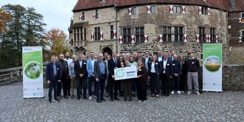 Eine Gruppe von ca. 40 Personen steht vor einem alten Steingebäude. Links und rechts der Gruppe stehen grüne Rollups. Drei Personen, die in der Mitte stehen, halten ein Schild mit der Aufschrift "Ökoprofit".
