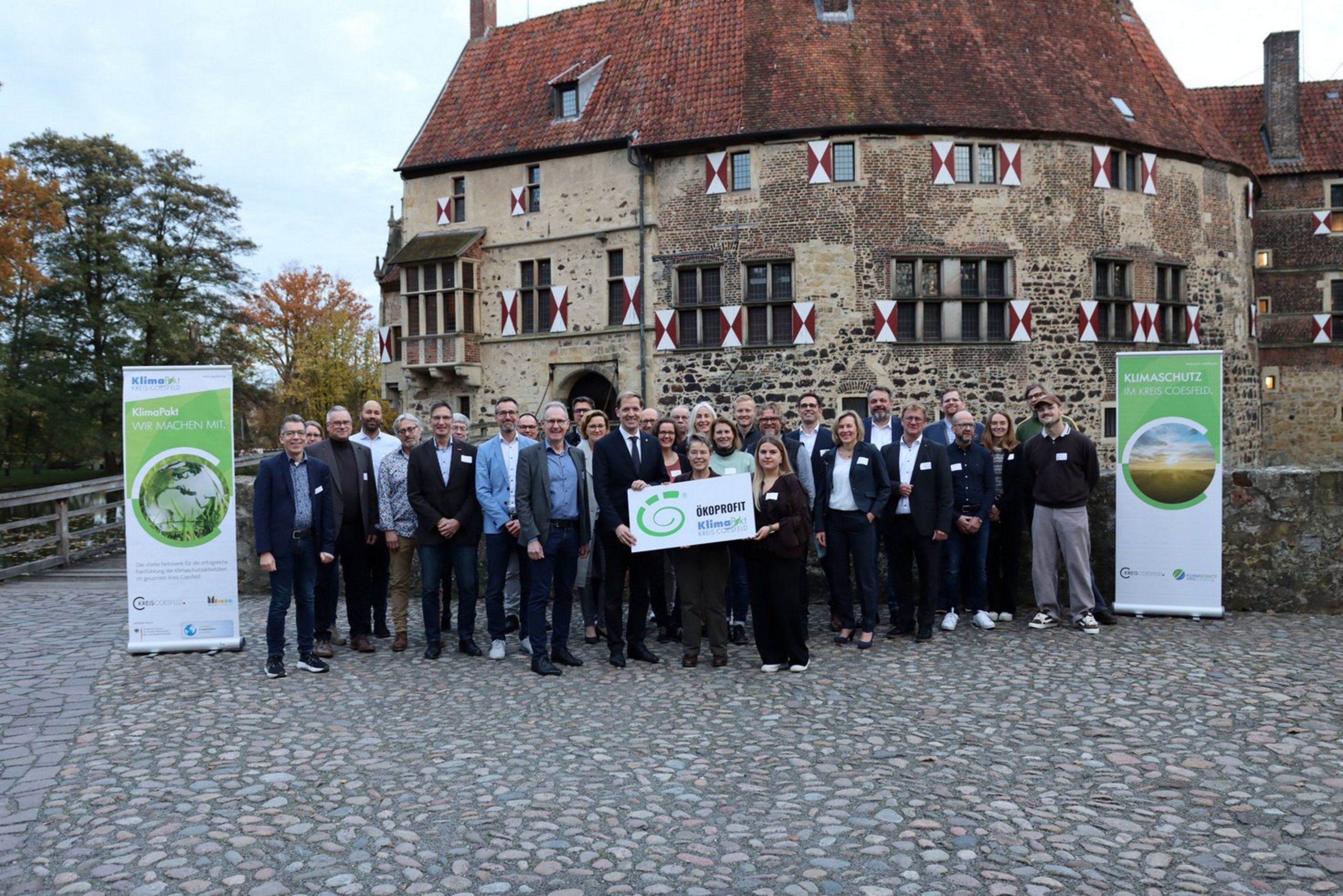 Eine Gruppe von ca. 40 Personen steht vor einem alten Steingebäude. Links und rechts der Gruppe stehen grüne Rollups. Drei Personen, die in der Mitte stehen, halten ein Schild mit der Aufschrift "Ökoprofit".