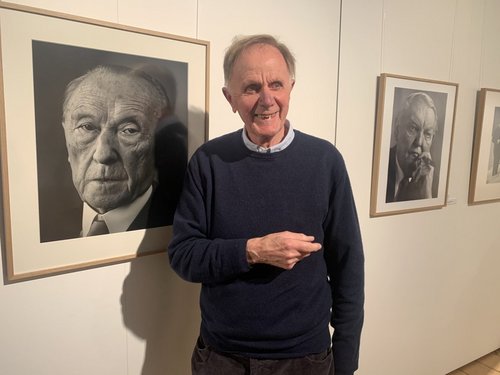 Ein älterer Mann in Pullover steht vor einer weißen Wand, an der zwei gerahmte  Fotografien in schwarz-weiß hängen. Sie zeigen Porträts.