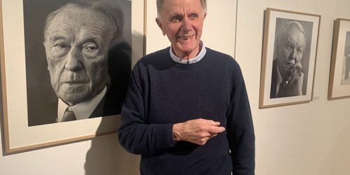 Ein älterer Mann in Pullover steht vor einer weißen Wand, an der zwei gerahmte  Fotografien in schwarz-weiß hängen. Sie zeigen Porträts.