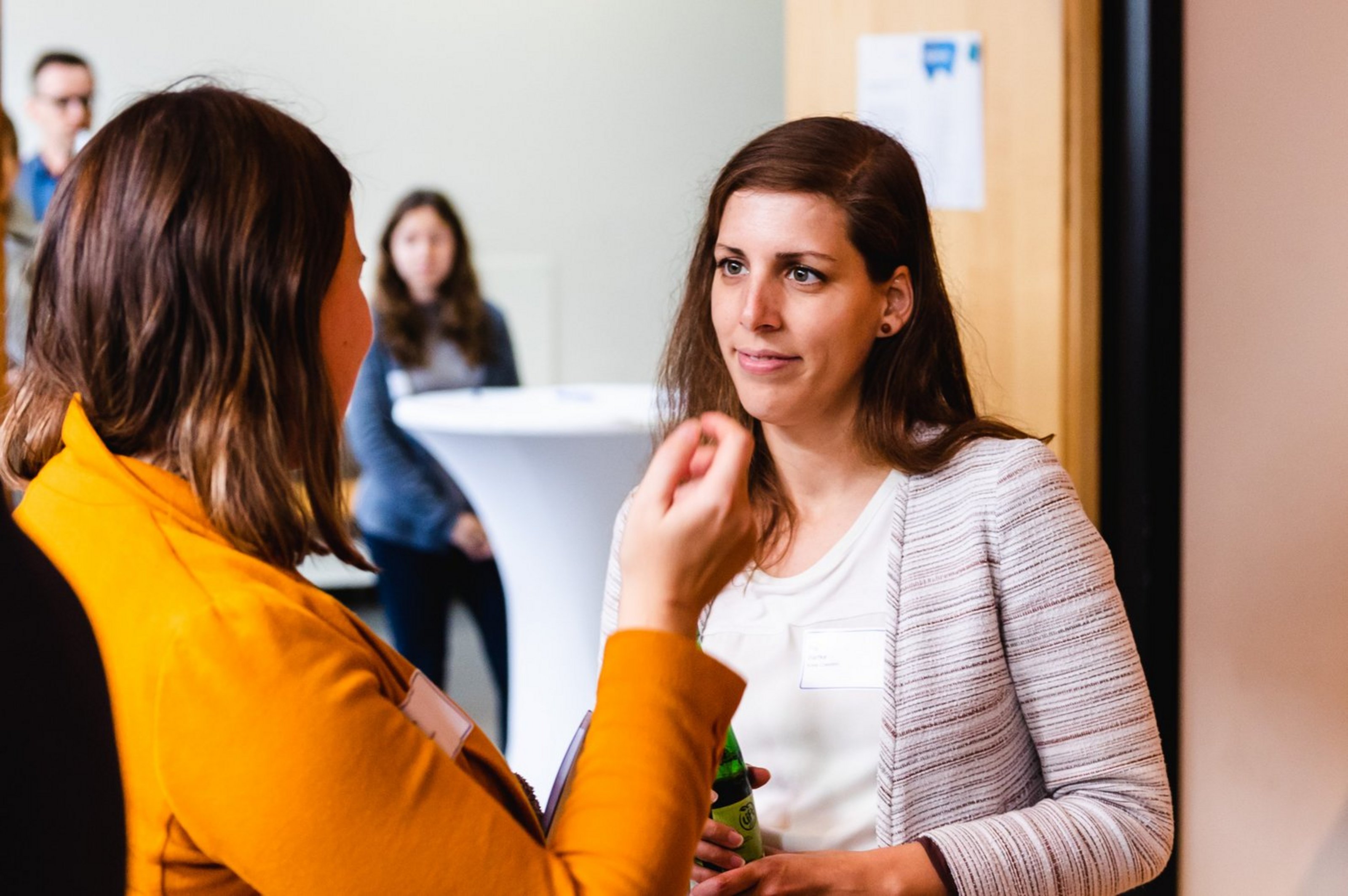 Zwei Frauen im Gespräch, eine trägt ein weißes Jackett, die andere ein orangefarbenes Oberteil, im Hintergrund unscharfe Personen und ein Stehtisch