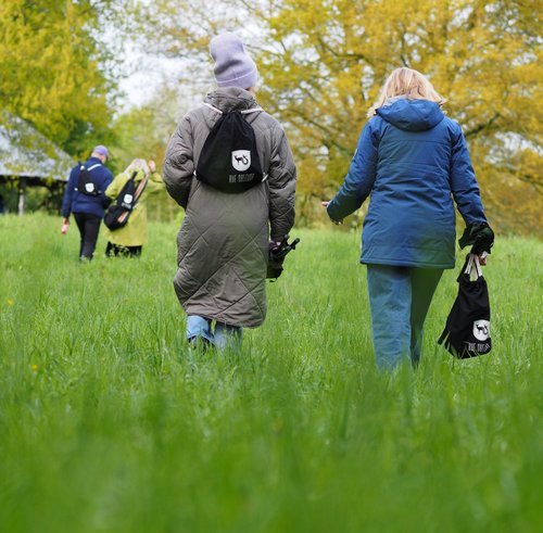 Zwei von hinten fotografierte Personen gehen nebeneinander durch kniehohes Gras. Im Hintergrund ist eine Baumreihe sowie eine alte Scheune zu sehen. 
