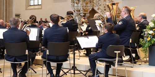 Man sieht eine Gruppe sitzender Musiker in dunklen Anzügen von hinten mit ihren Instrumenten. Sie sind in einer Kirche. Im Hintergrund sieht man das Publikum.