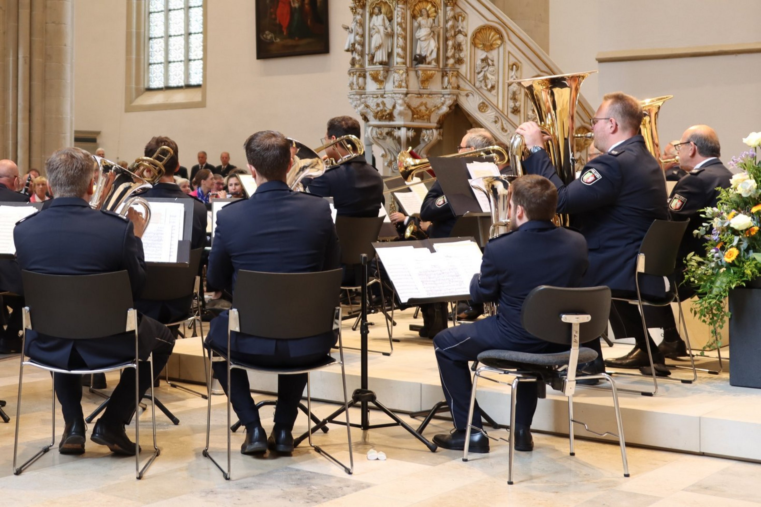 Man sieht eine Gruppe sitzender Musiker in dunklen Anzügen von hinten mit ihren Instrumenten. Sie sind in einer Kirche. Im Hintergrund sieht man das Publikum.