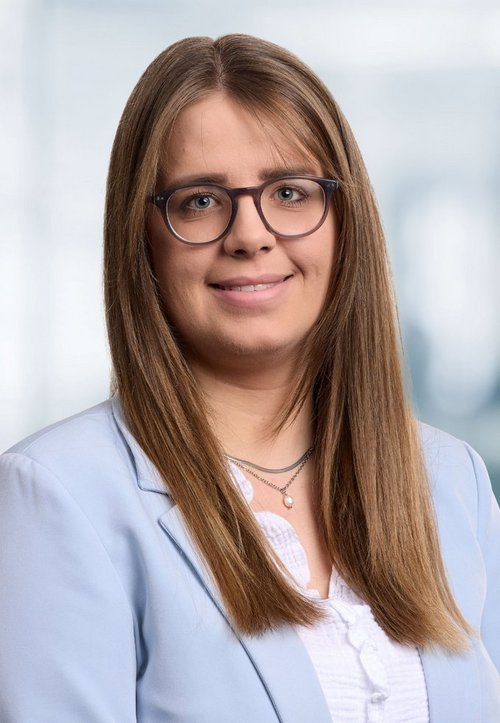 Eine Frau mit Blazer und Brille lächelt in die Kamera. Der Hintergrund ist blau.