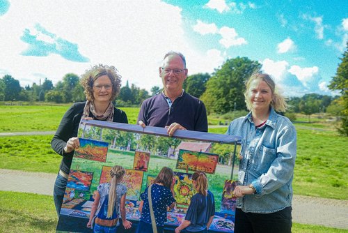 Drei Personen halten gemeinsam ein Plakat mit bunten Bildern und freuen sich auf „Kunst im Park“ in Lüdinghausen.