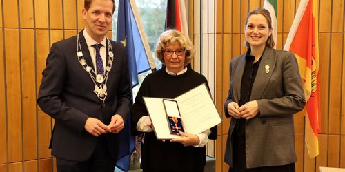 Drei Personen stehen vor vier Fahnen. Die Frau in der Mitte hält eine Urkunde und ein Kreuz in einer Schatulle in die Kamera.