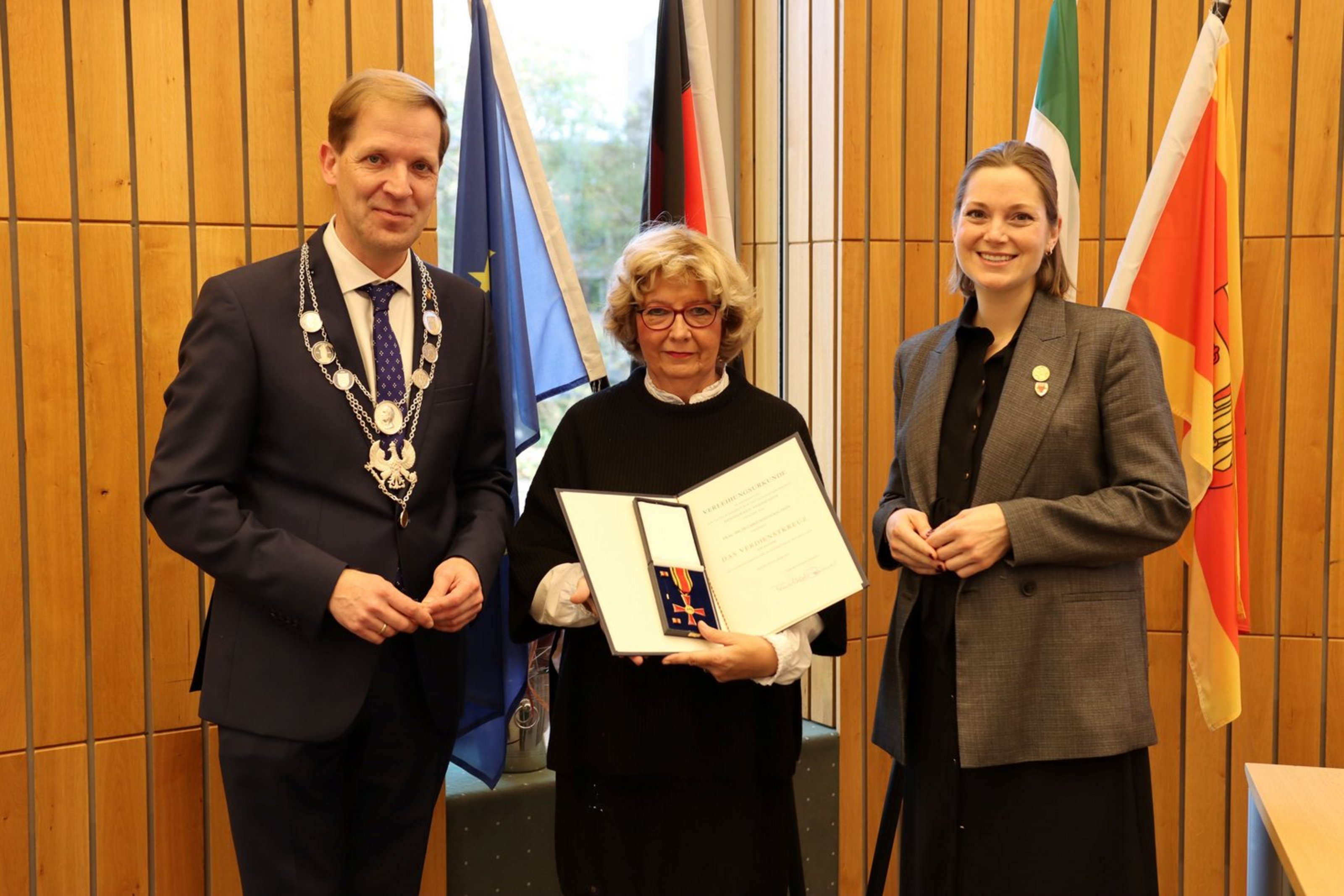 Drei Personen stehen vor vier Fahnen. Die Frau in der Mitte hält eine Urkunde und ein Kreuz in einer Schatulle in die Kamera.