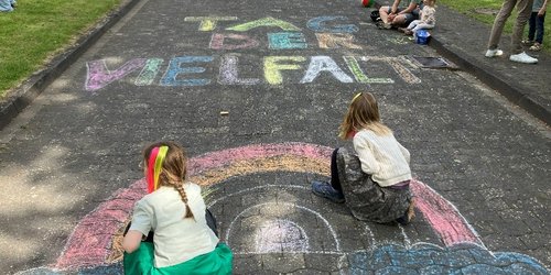 Auf eienr Straße ist mit Kreide ein Regenbogen und die Aufschrift "Tag der Vielfalt" geschrieben. Zwei Kinder sitzen auf dem Regenbogen.