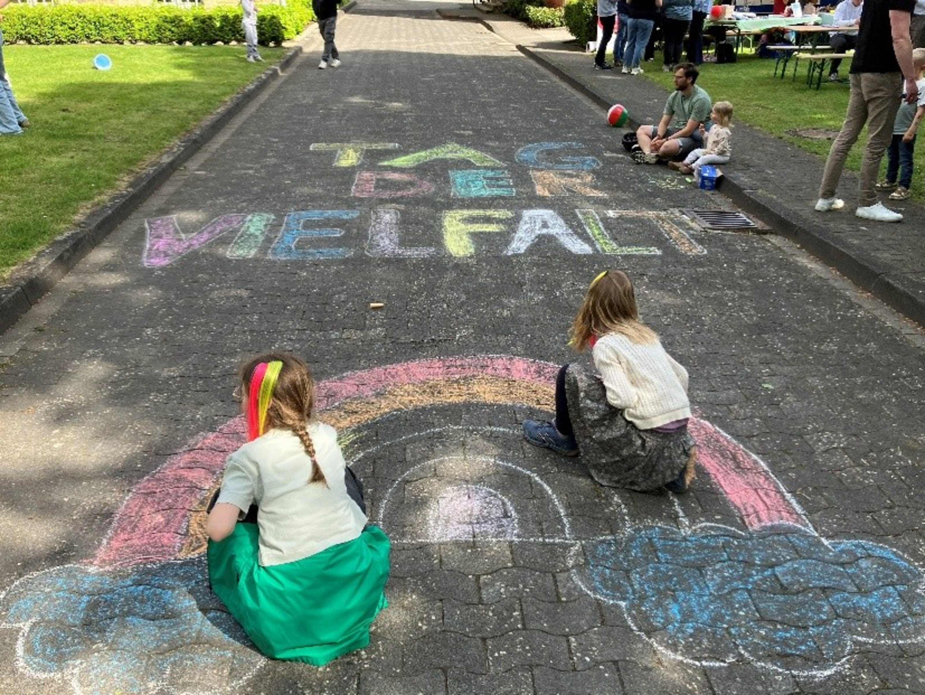 Auf eienr Straße ist mit Kreide ein Regenbogen und die Aufschrift "Tag der Vielfalt" geschrieben. Zwei Kinder sitzen auf dem Regenbogen.