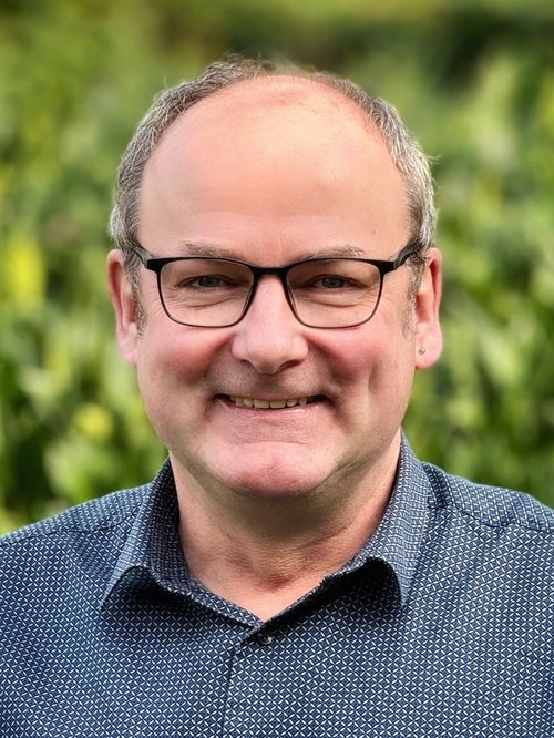 Eine Mann mit Brille und grauem Hemd lächelt in die Kamera. Der Hintergrund ist grün.