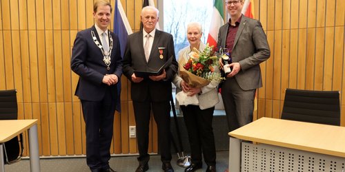 In einem holzvertäfelten Raum stehen vier Personen nebeneinander. Die drei Männer tragen Anzüge, die Frau hält einen Blumenstraß.