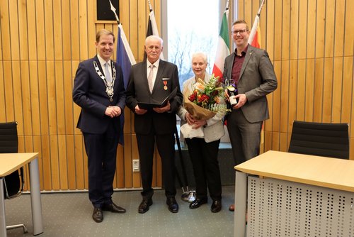 In einem holzvertäfelten Raum stehen vier Personen nebeneinander. Die drei Männer tragen Anzüge, die Frau hält einen Blumenstraß.