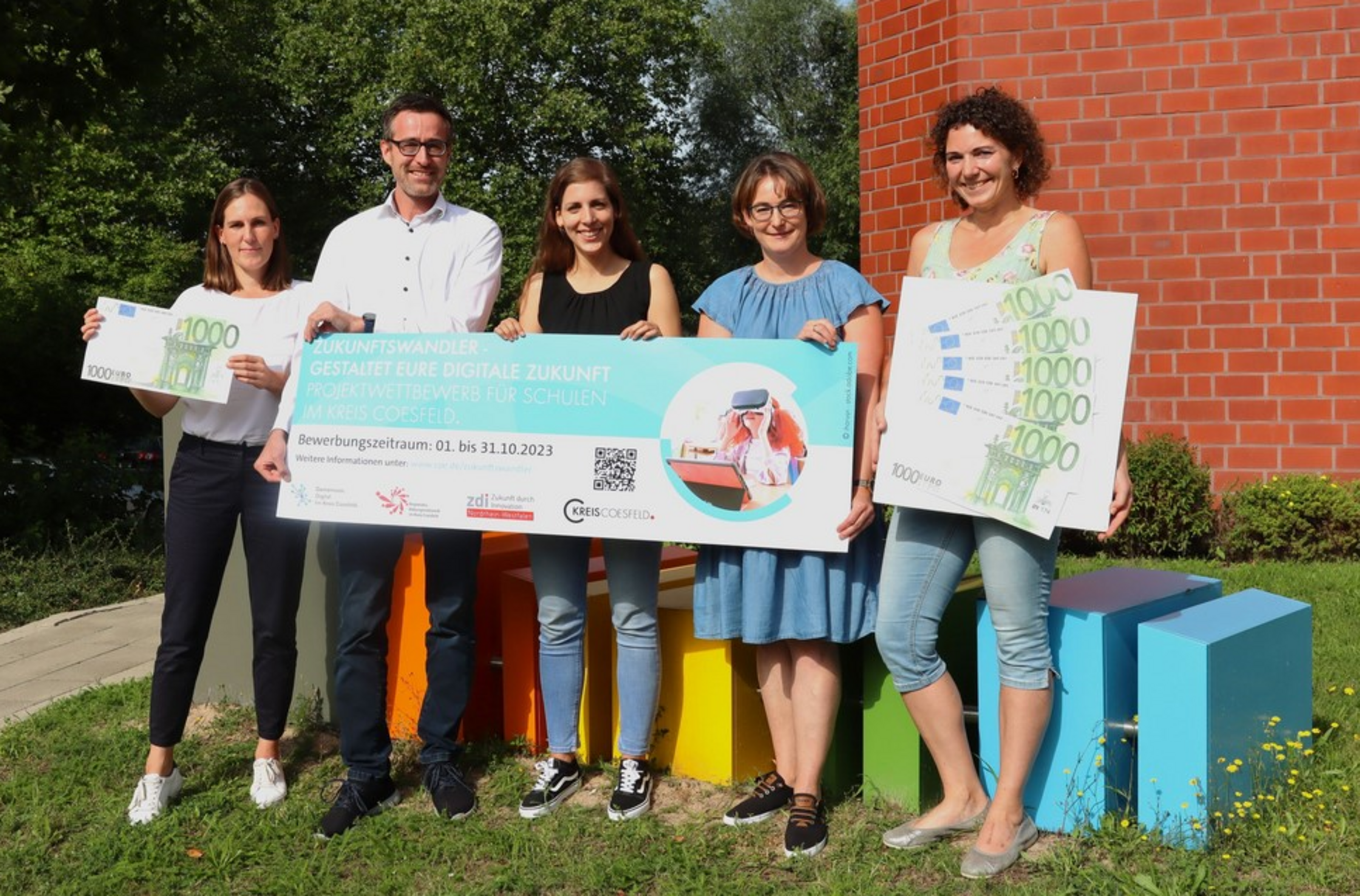 Fünf Personen stehen im Freien vor bunten Sitzwürfeln und halten große Schecks mit der Aufschrift '1000 Euro' und einem Banner mit Text und einem Bild einer Person am Laptop