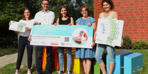 Fünf Personen stehen im Freien vor bunten Sitzwürfeln und halten große Schecks mit der Aufschrift '1000 Euro' und einem Banner mit Text und einem Bild einer Person am Laptop