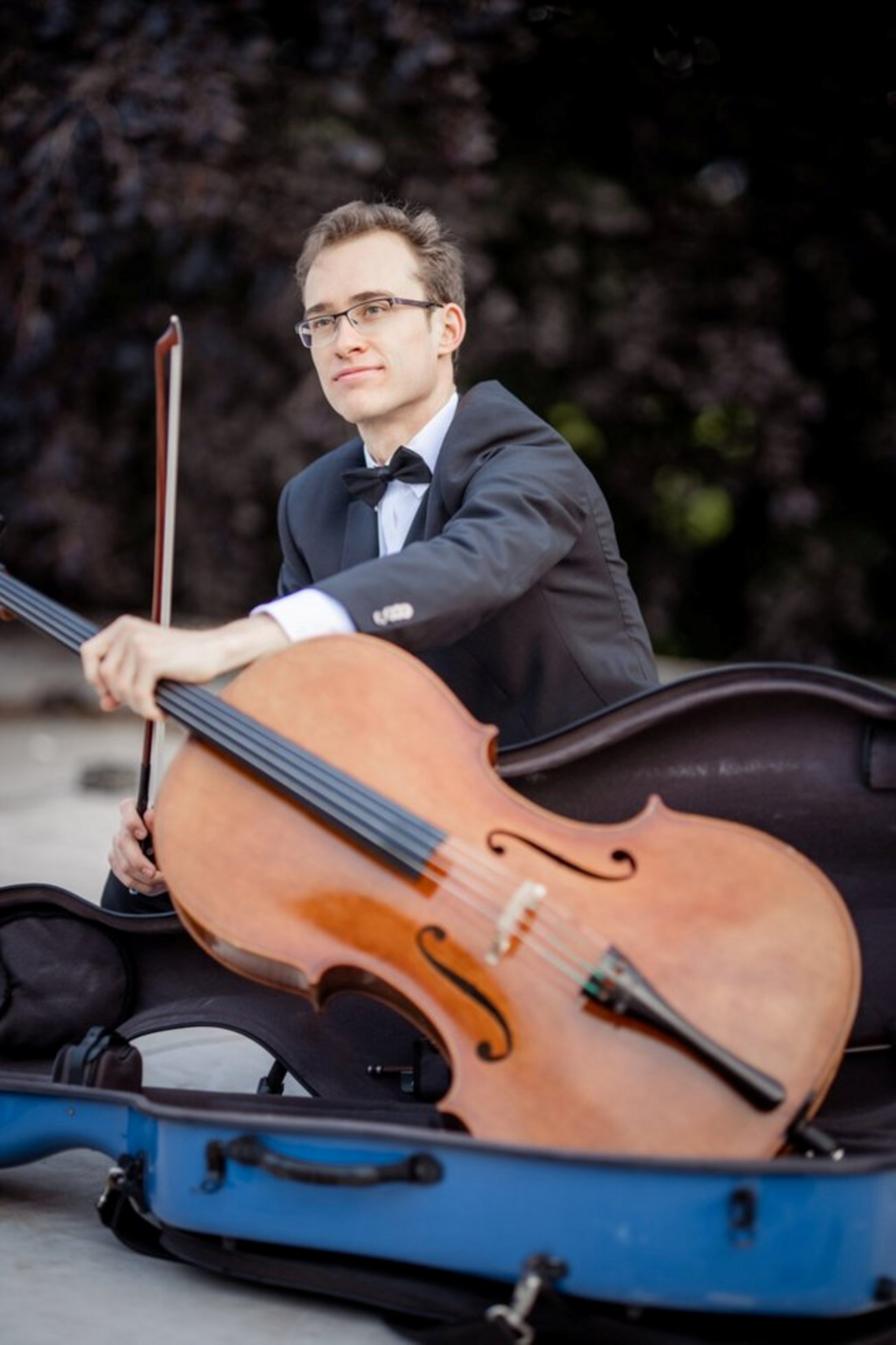 Ein Mann im Anzug hält ein Cello in der Hand sowie einen Bogen. Das Cello liegt in einem schwarzen Cello-Koffer. 
