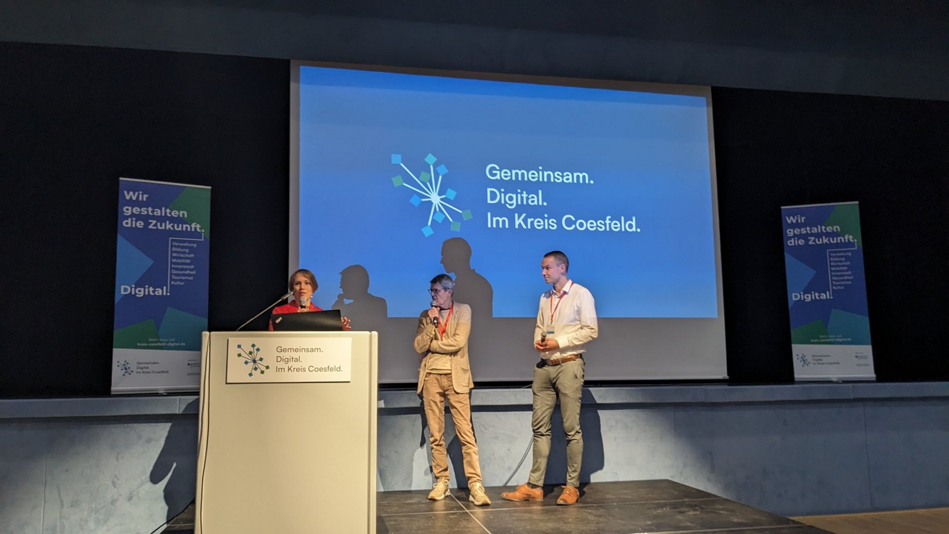 Drei Personen stehen auf einer Bühne vor einer Leinwand mit der Projektion 'Gemeinsam. Digital. Im Kreis Coesfeld.' und zwei Bannern mit dem Text 'Wir gestalten die Zukunft. Digital!'